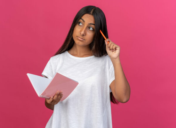 jeune fille t shirt blanc tenant cahier crayon regardant cote perplexe se gratter tete crayon pensant debout fond rose scaled